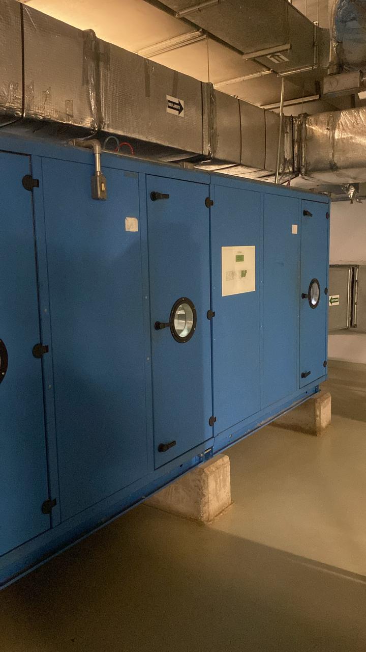 Imagem de uma unidade de tratamento de ar (UTA) grande e de cor azul instalada em um ambiente técnico. A UTA está apoiada em blocos de concreto e conectada a dutos de ventilação isolados que distribuem o ar para várias áreas. Na parte superior do equipamento, há uma seta indicando a direção do fluxo de ar.